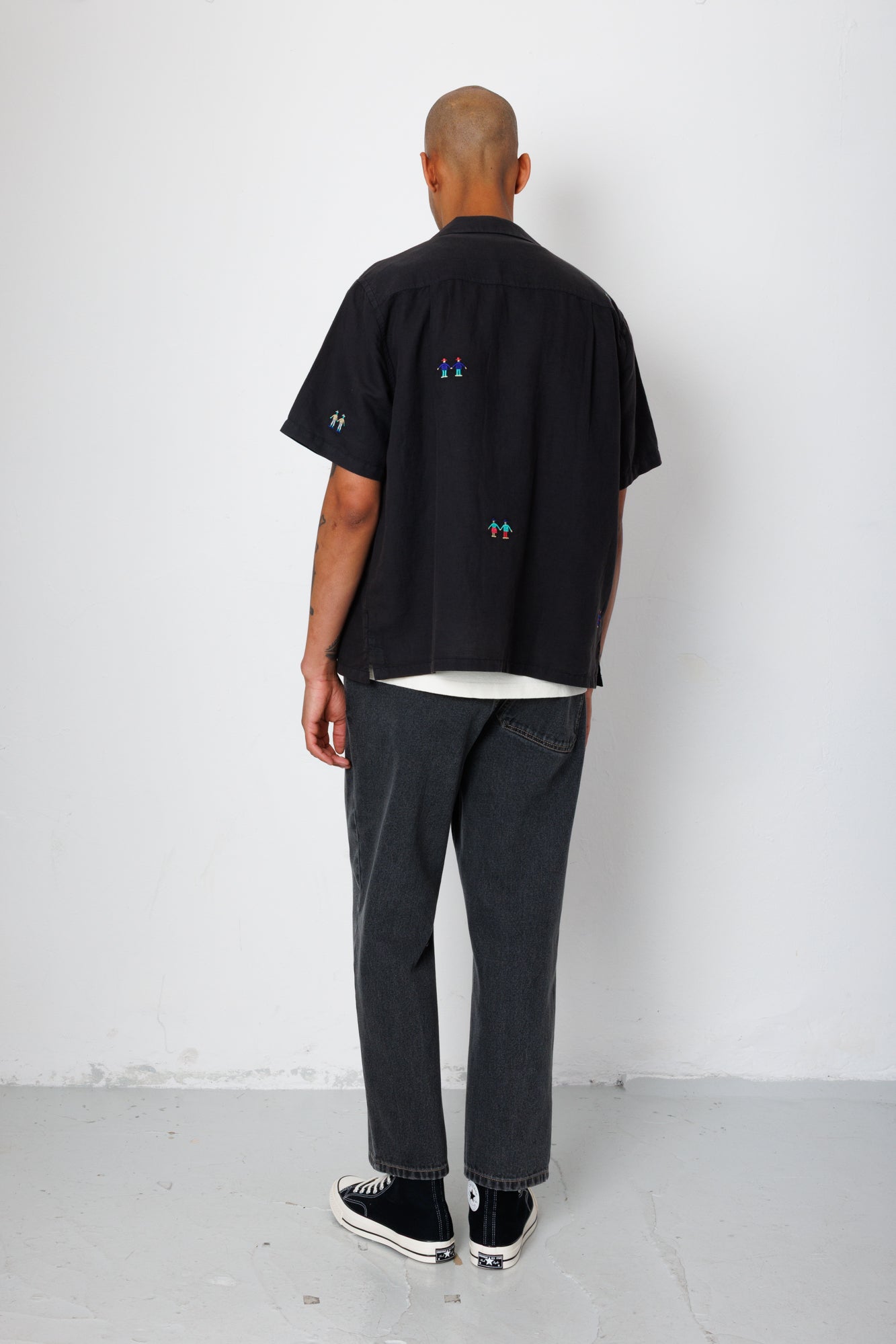 Model facing a white wall while wearing a black shirt with embroidery.