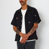 A man wearing a black open button down shirt with embroidery detail and black jeans.