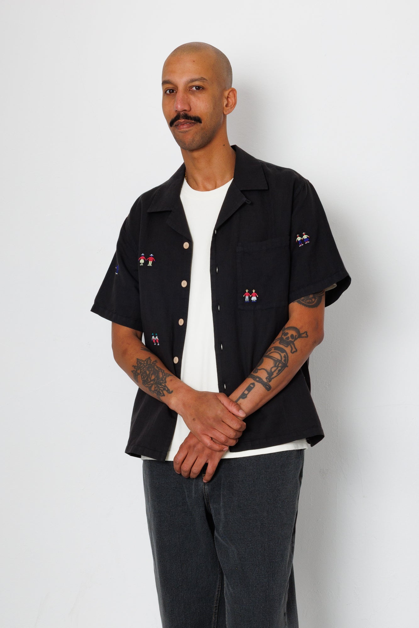 A man wearing a black open button down shirt with embroidery detail and black jeans.