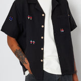 Close up of a man wearing a black open button down shirt with embroidery detail and black jeans.