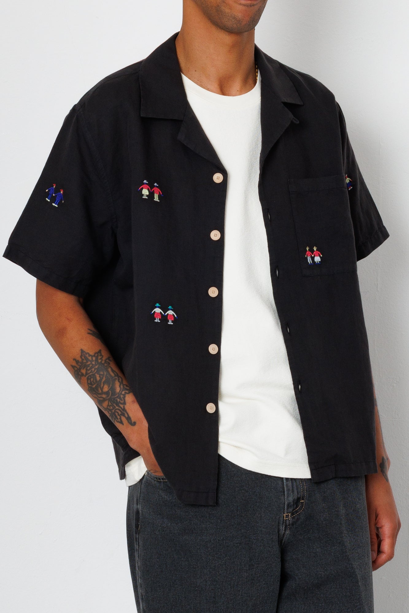 Close up of a man wearing a black open button down shirt with embroidery detail and black jeans.