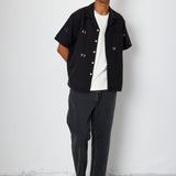 A man wearing a black open button down shirt with embroidery detail and black jeans.