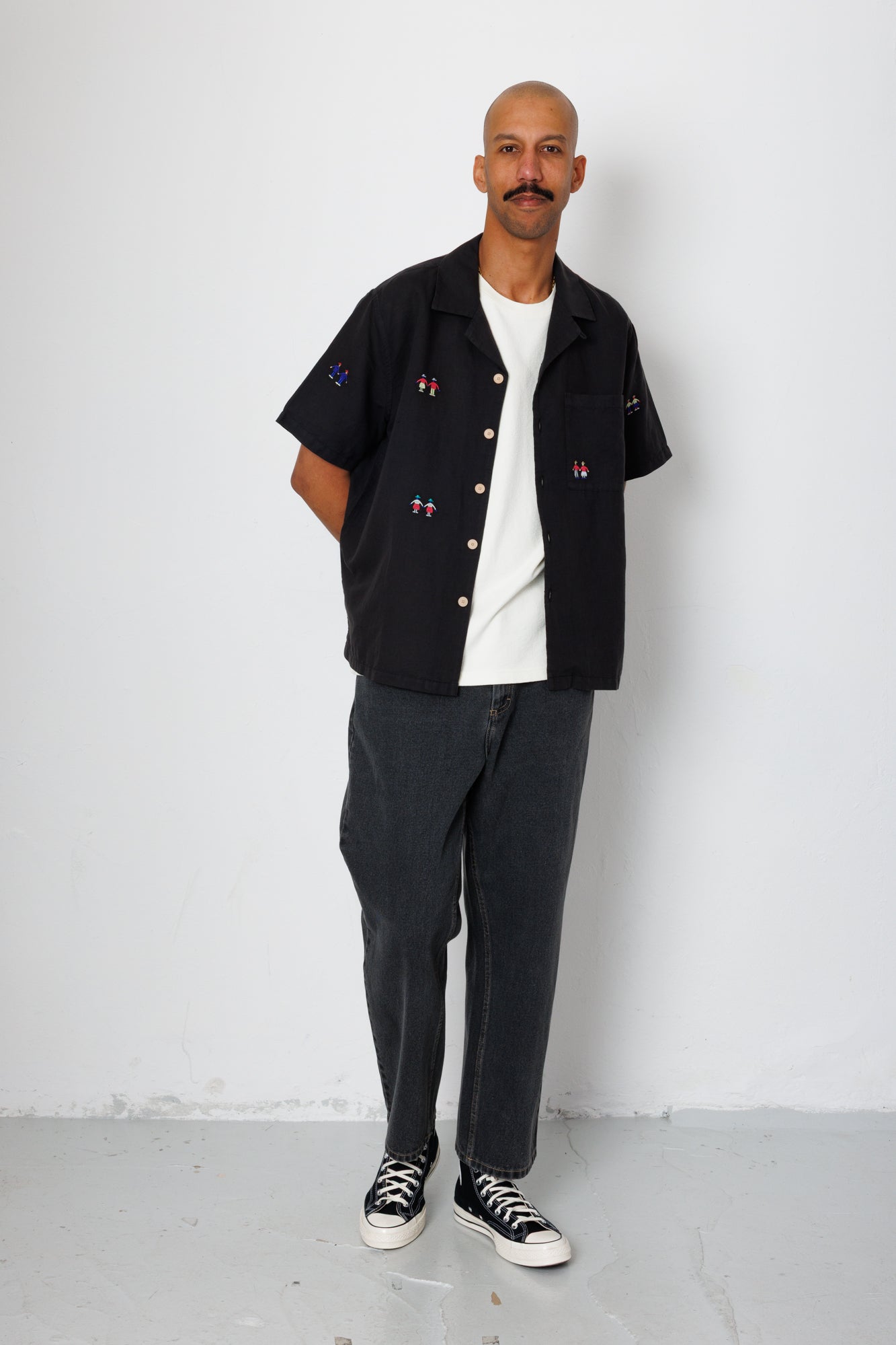 A man wearing a black open button down shirt with embroidery detail and black jeans.