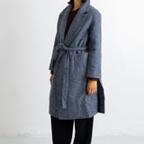 A woman modeling a long gray wool-blend coat with a tie belt, worn over a dark top and black trousers, standing against a plain white studio background.