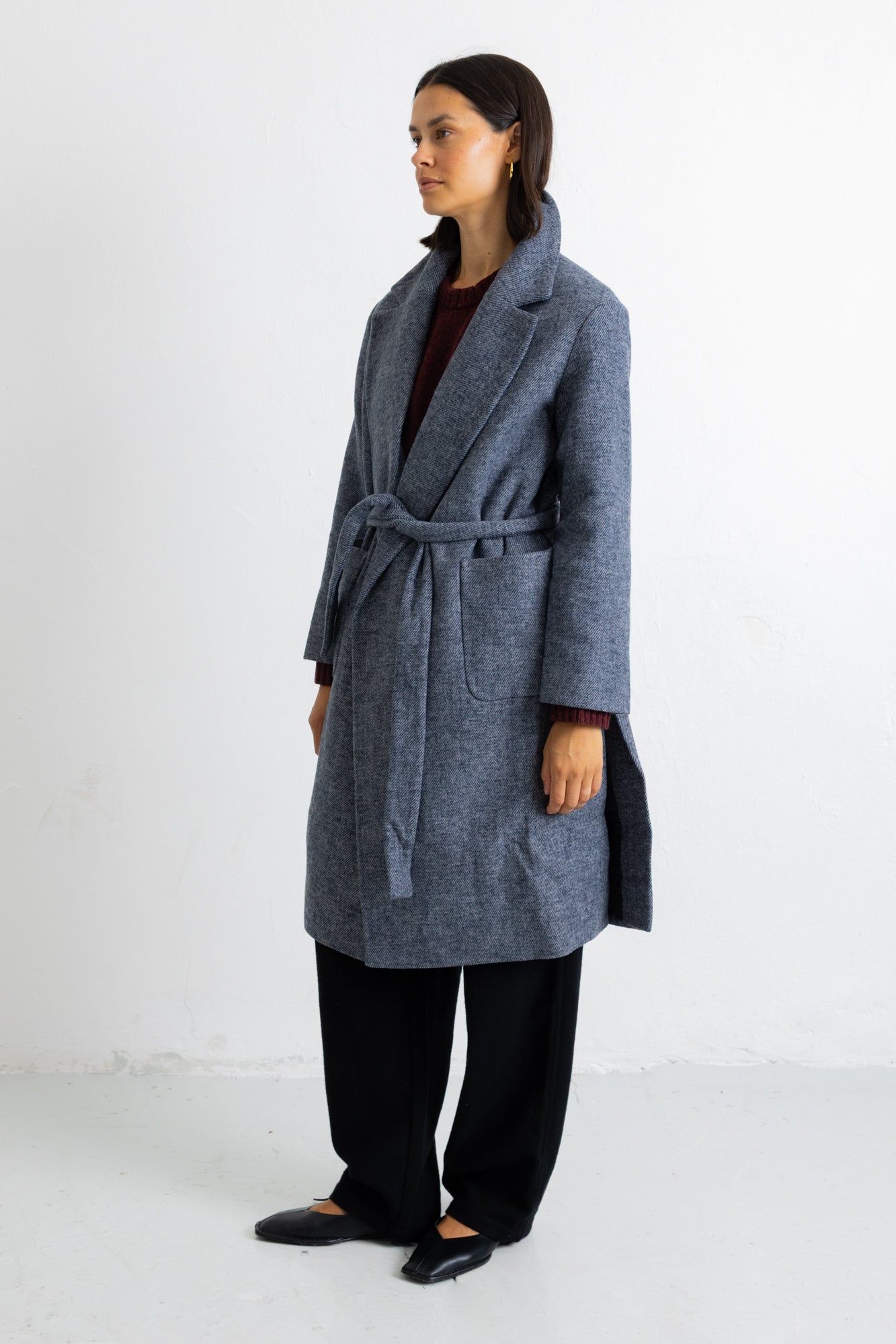A woman modeling a long gray wool-blend coat with a tie belt, worn over a dark top and black trousers, standing against a plain white studio background.