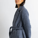 A woman modeling a long, open-front wool coat in a heathered slate blue-gray color, styled with a dark burgundy top, black wide-leg trousers, and black flats, set against a plain white studio background.