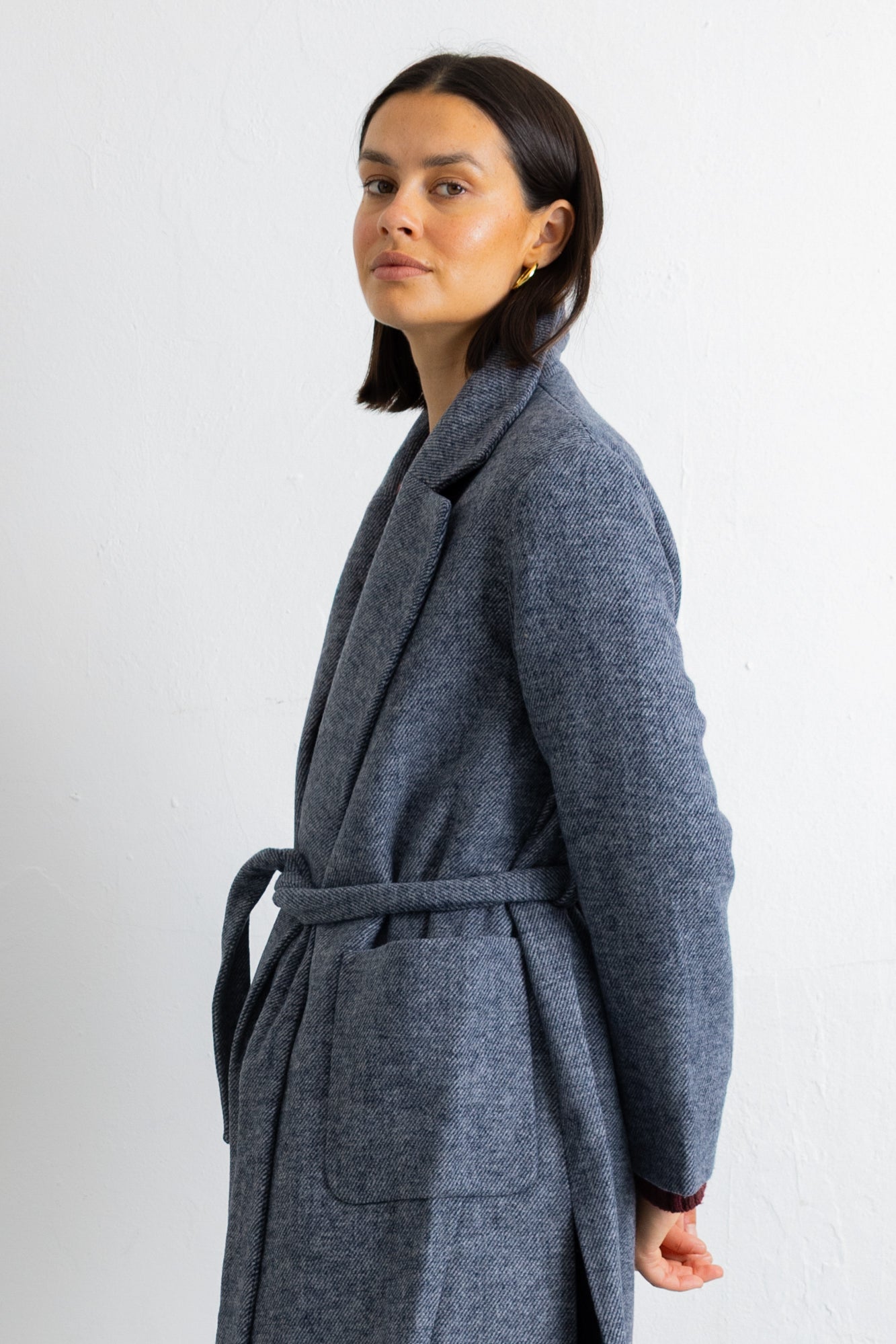 A woman modeling a long, open-front wool coat in a heathered slate blue-gray color, styled with a dark burgundy top, black wide-leg trousers, and black flats, set against a plain white studio background.