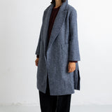 A woman modeling a long, open-front wool coat in a heathered slate blue-gray color, styled with a dark burgundy top, black wide-leg trousers, and black flats, set against a plain white studio background.
