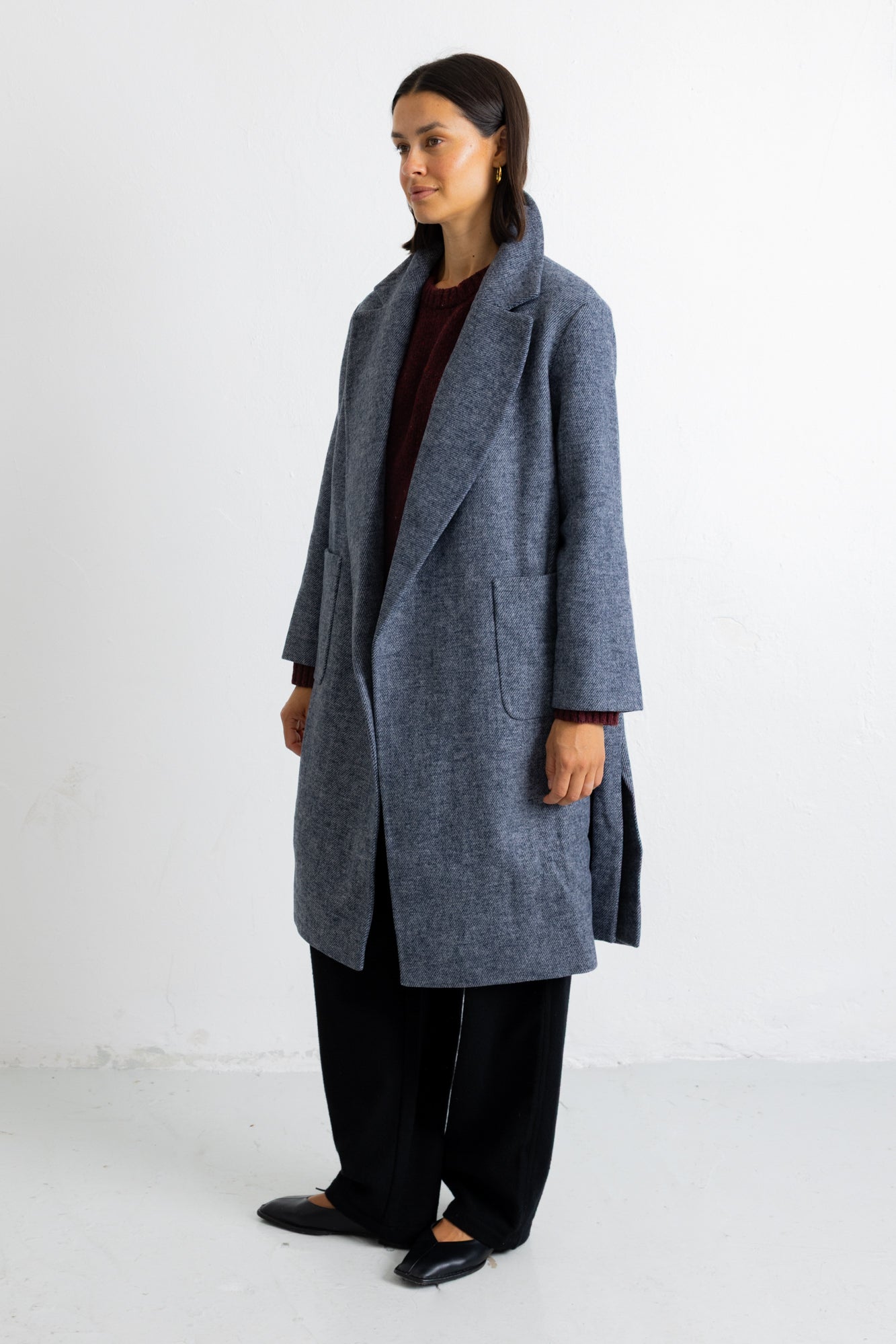A woman modeling a long, open-front wool coat in a heathered slate blue-gray color, styled with a dark burgundy top, black wide-leg trousers, and black flats, set against a plain white studio background.