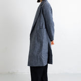 A woman stands in profile wearing a long charcoal gray textured coat over black wide-leg trousers and black flats, photographed against a plain white studio background.
