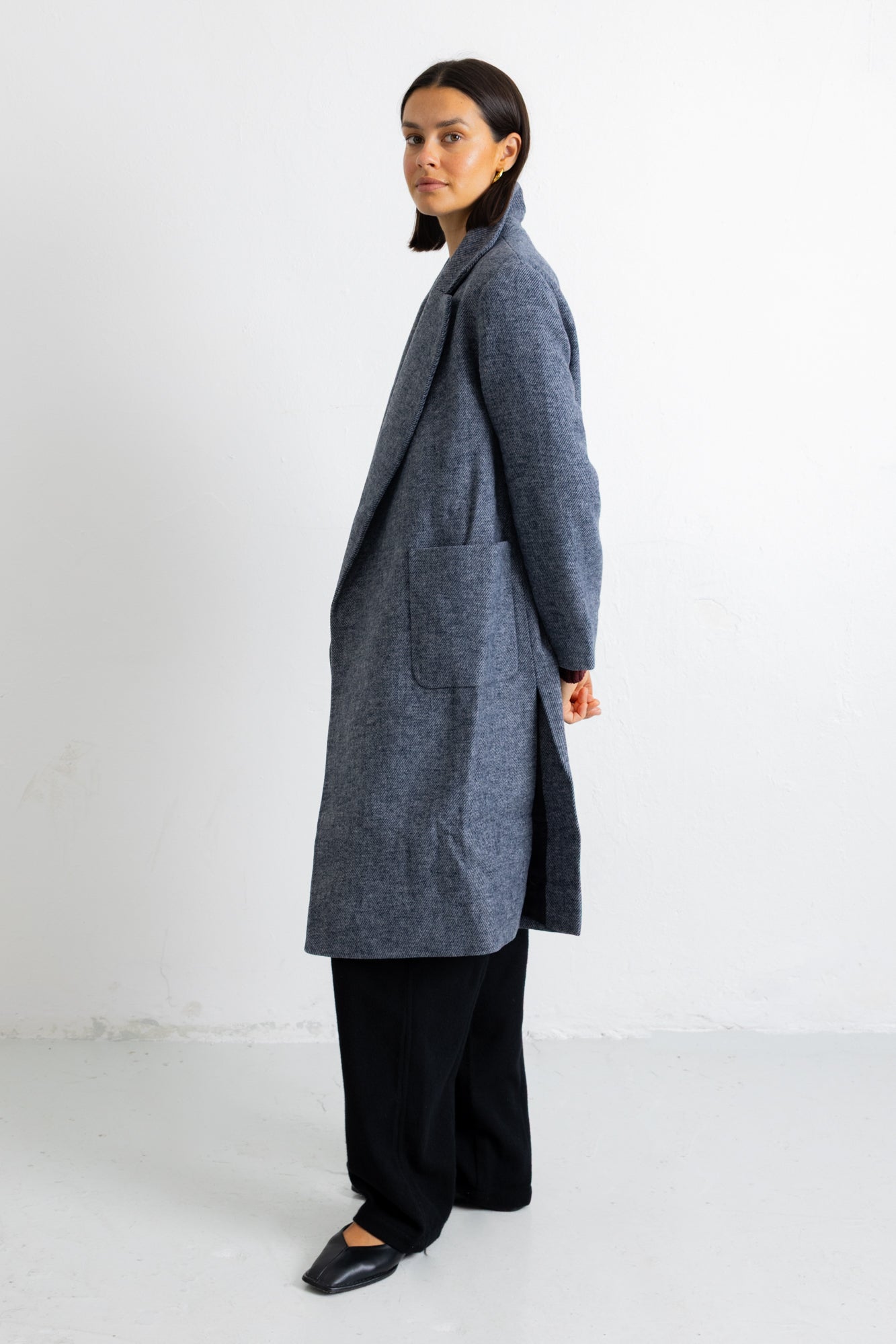 A woman stands in profile wearing a long charcoal gray textured coat over black wide-leg trousers and black flats, photographed against a plain white studio background.
