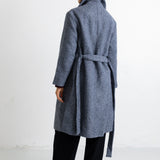 A woman modeling a long, open-front wool coat in a heathered slate blue-gray color, styled with a dark burgundy top, black wide-leg trousers, and black flats, set against a plain white studio background.