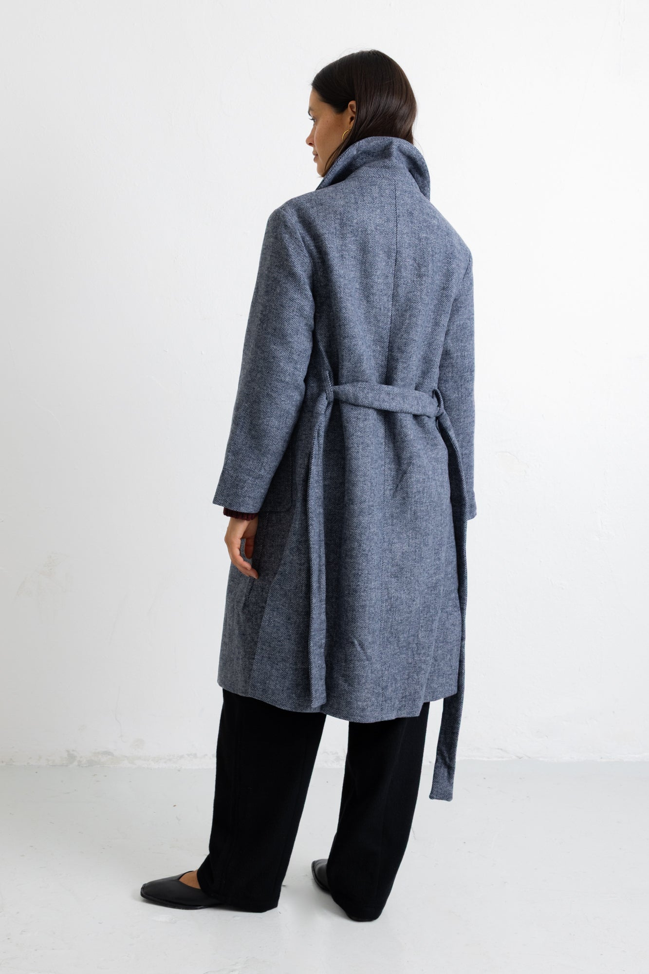 A woman modeling a long, open-front wool coat in a heathered slate blue-gray color, styled with a dark burgundy top, black wide-leg trousers, and black flats, set against a plain white studio background.