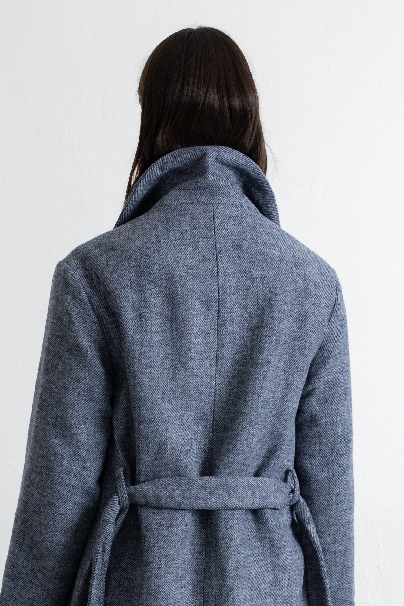 A person shown from the back wearing a textured blue-gray wrap coat with a high collar and waist tie, against a plain white background.