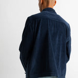 A man viewed from behind wearing a dark navy blue corduroy jacket over a light blue shirt and green pants, standing against a plain white wall.