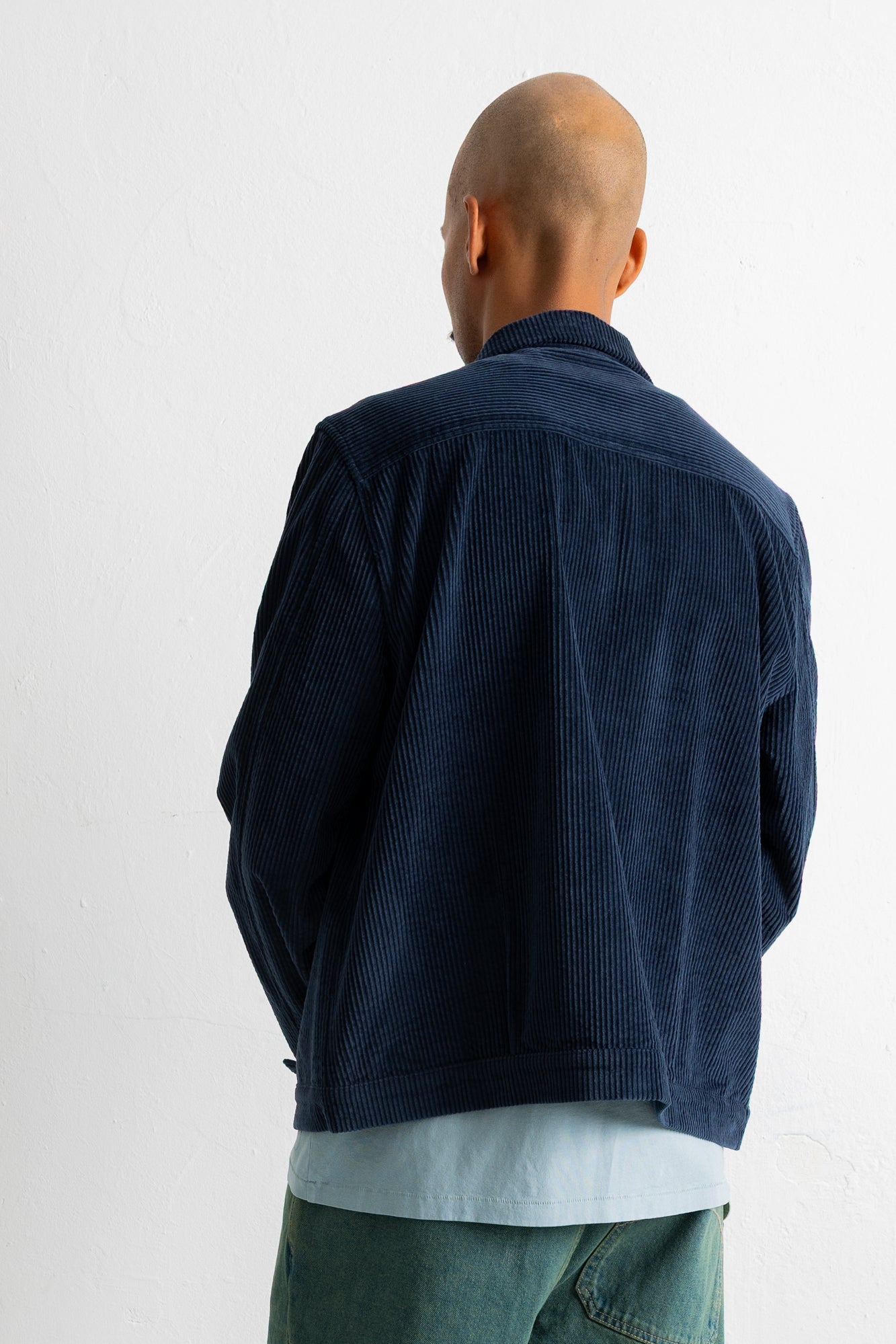 A man viewed from behind wearing a dark navy blue corduroy jacket over a light blue shirt and green pants, standing against a plain white wall.