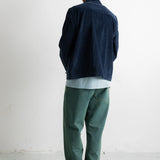 A man viewed from behind wearing a dark navy corduroy jacket, a light blue t-shirt, green trousers, and black-and-white sneakers, standing against a plain white wall.