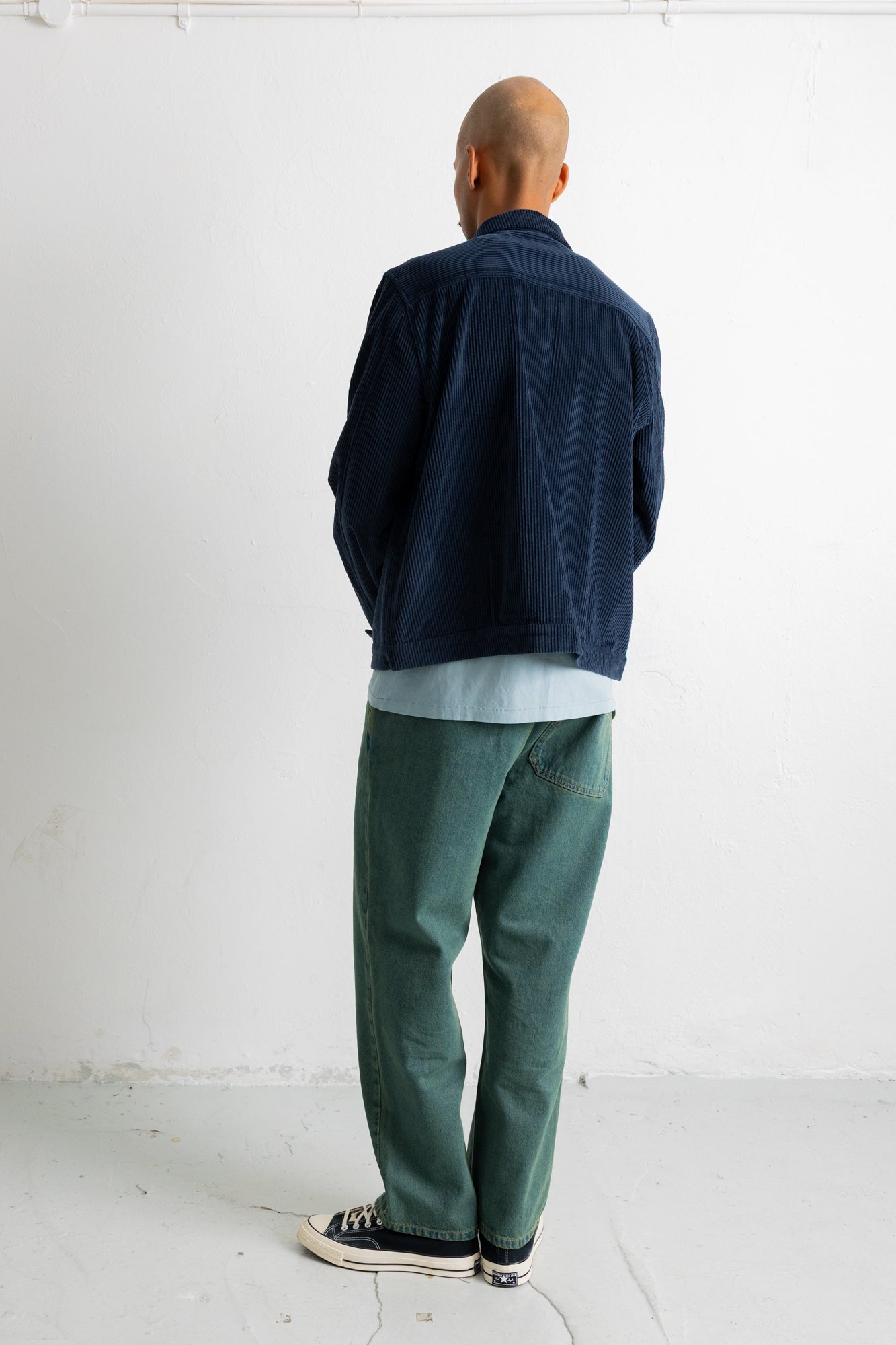 A man viewed from behind wearing a dark navy corduroy jacket, a light blue t-shirt, green trousers, and black-and-white sneakers, standing against a plain white wall.