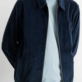 A man wearing a dark navy blue corduroy zip-up jacket over a light blue t-shirt and green jeans, shown from the neck down against a white wall.