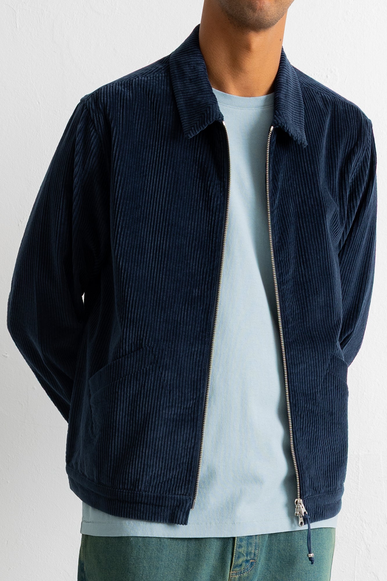 A man wearing a dark navy blue corduroy zip-up jacket over a light blue t-shirt and green jeans, shown from the neck down against a white wall.