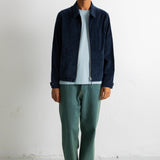 A man wearing a dark navy blue zip-up jacket over a light blue t-shirt, paired with green trousers and black-and-white sneakers, standing against a white wall.