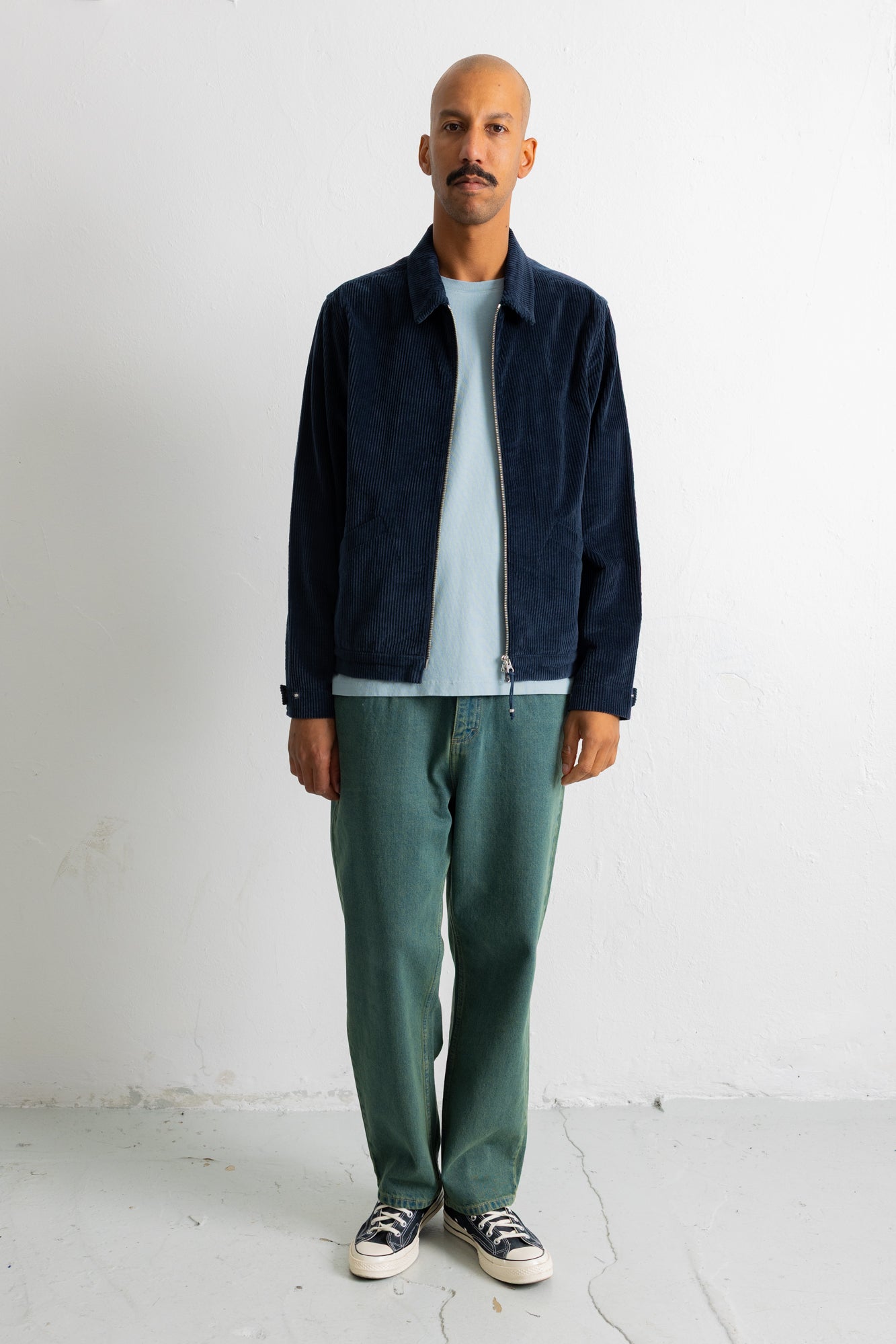 A man wearing a dark navy blue zip-up jacket over a light blue t-shirt, paired with green trousers and black-and-white sneakers, standing against a white wall.