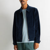 A man wearing a dark navy blue corduroy jacket over a light blue t-shirt and teal pants, standing against a white wall.