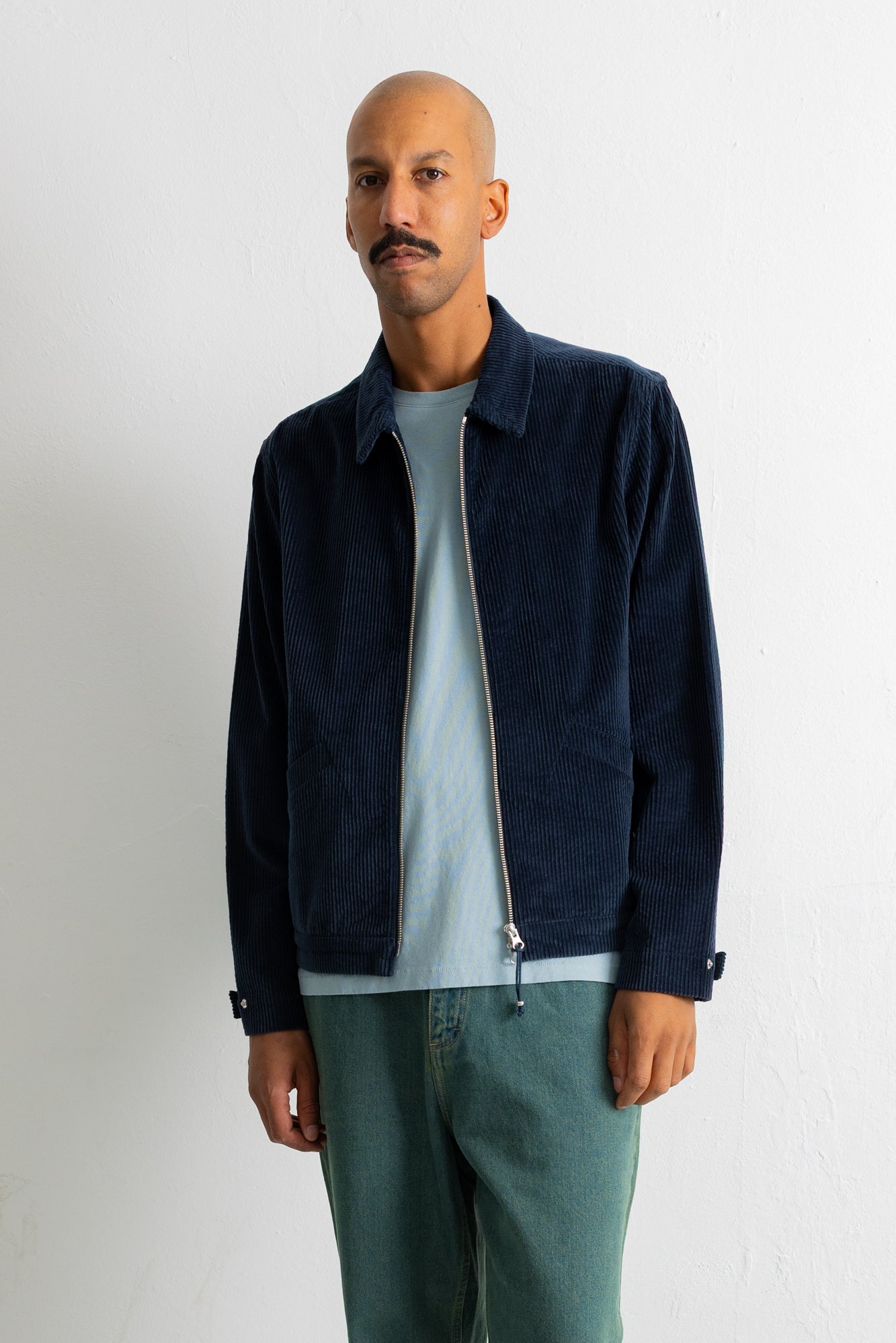 A man wearing a dark navy blue corduroy jacket over a light blue t-shirt and teal pants, standing against a white wall.