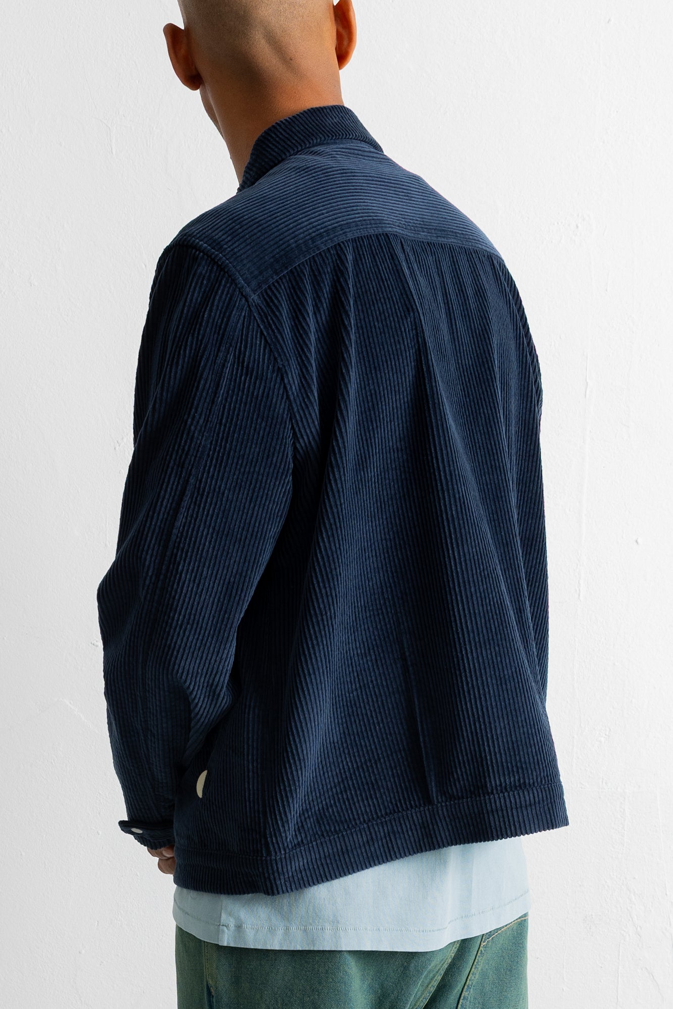 A man wearing a dark navy blue corduroy jacket, shown from a rear three-quarter view against a plain white wall.
