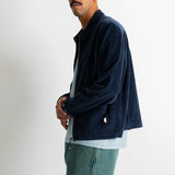 A man wearing a dark navy corduroy jacket over a light blue shirt and teal jeans, standing against a plain white wall.
