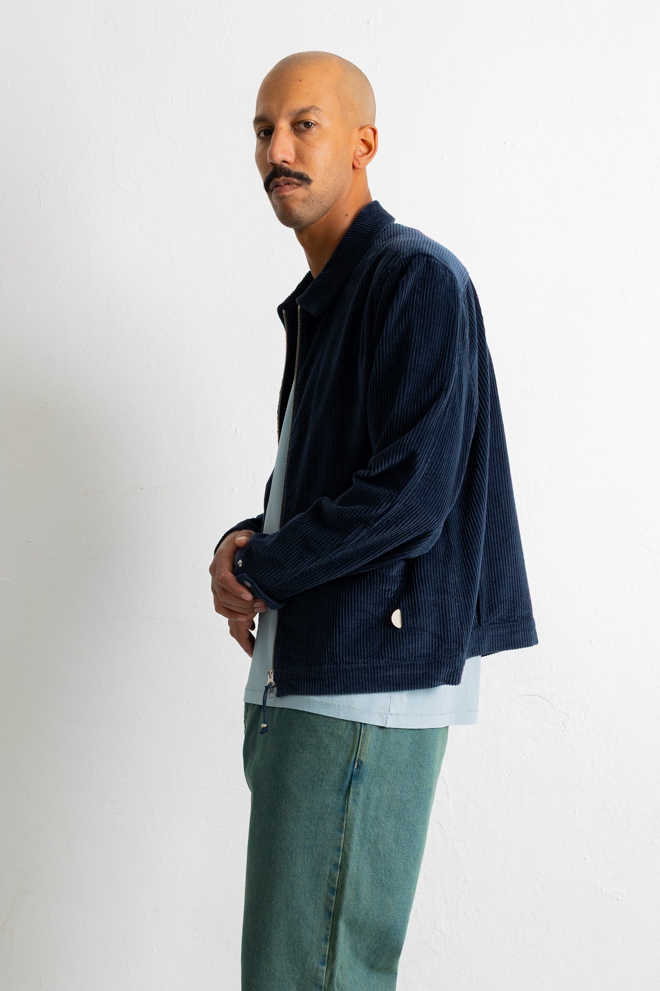 A man wearing a dark navy corduroy jacket over a light blue shirt and teal jeans, standing against a plain white wall.