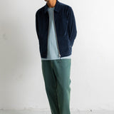 A man wearing a navy blue corduroy zip-up jacket over a light blue t-shirt, paired with green trousers and black-and-white sneakers, standing against a white wall.