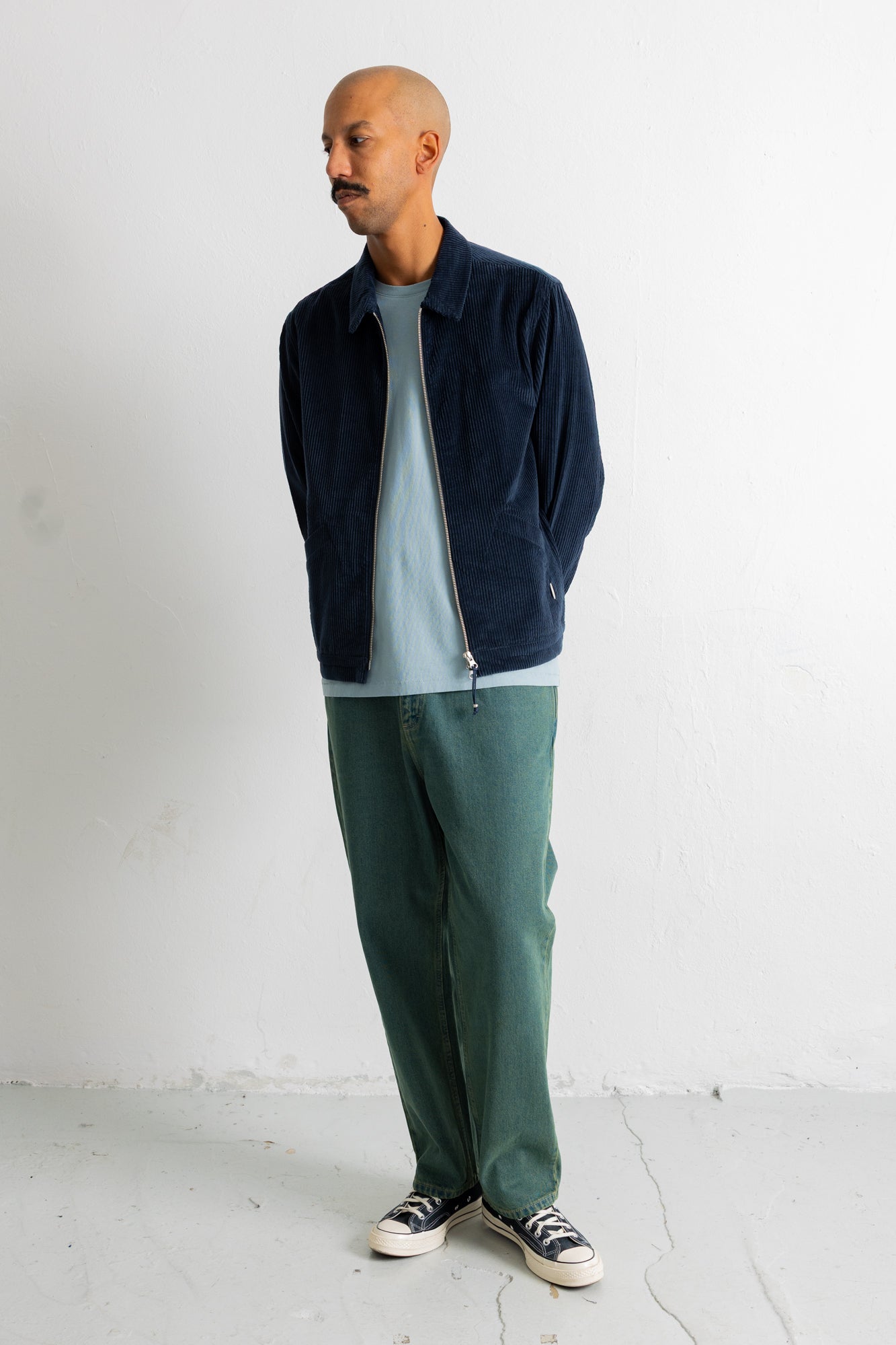 A man wearing a navy blue corduroy zip-up jacket over a light blue t-shirt, paired with green trousers and black-and-white sneakers, standing against a white wall.