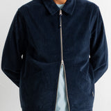 A man wearing a dark navy blue corduroy zip-up jacket over a light blue t-shirt and green jeans, photographed against a white wall.