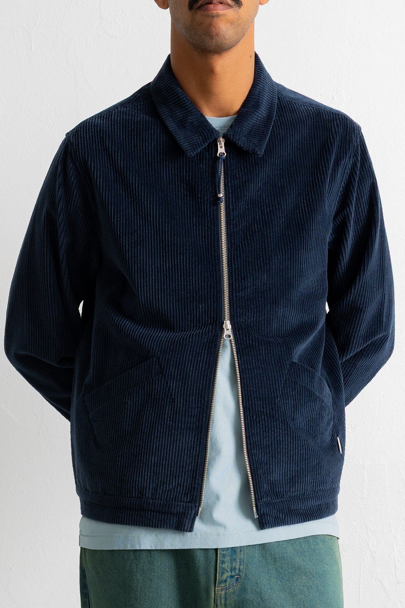A man wearing a dark navy blue corduroy zip-up jacket over a light blue t-shirt and green jeans, photographed against a white wall.