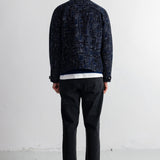 A man viewed from behind wearing a dark navy blue patterned jacket, black jeans, and black shoes, standing against a plain white wall.