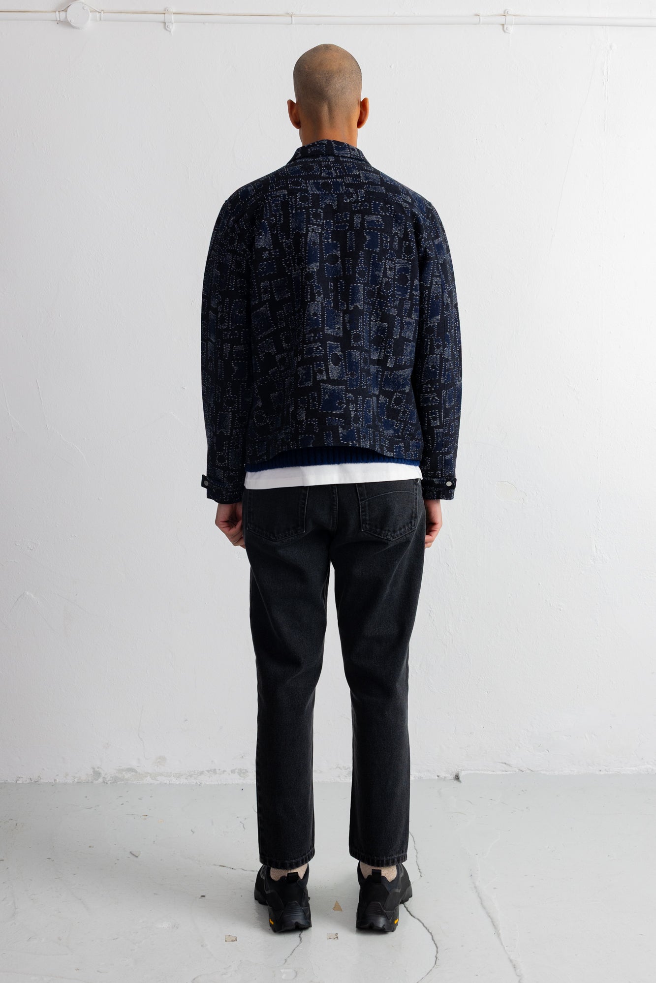 A man viewed from behind wearing a dark navy blue patterned jacket, black jeans, and black shoes, standing against a plain white wall.