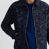 A man wearing a dark navy patterned zip-up jacket over a blue knit sweater and dark jeans, shown from the chin down against a plain white wall.