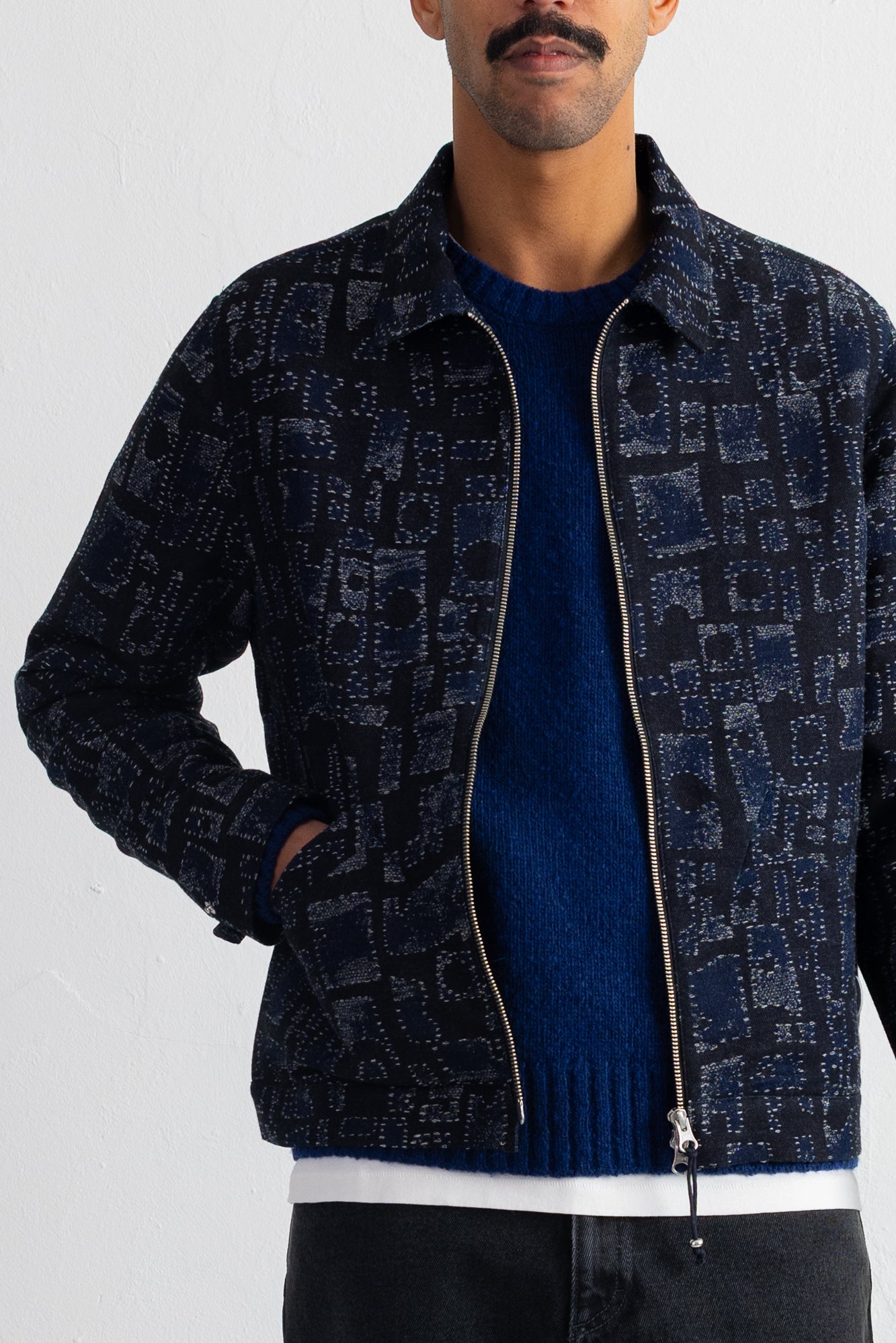 A man wearing a dark navy patterned zip-up jacket over a blue knit sweater and dark jeans, shown from the chin down against a plain white wall.