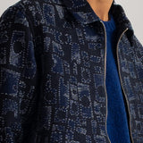 Close-up of a man wearing a dark navy patterned zip-up jacket over a blue knit sweater, against a light gray background.