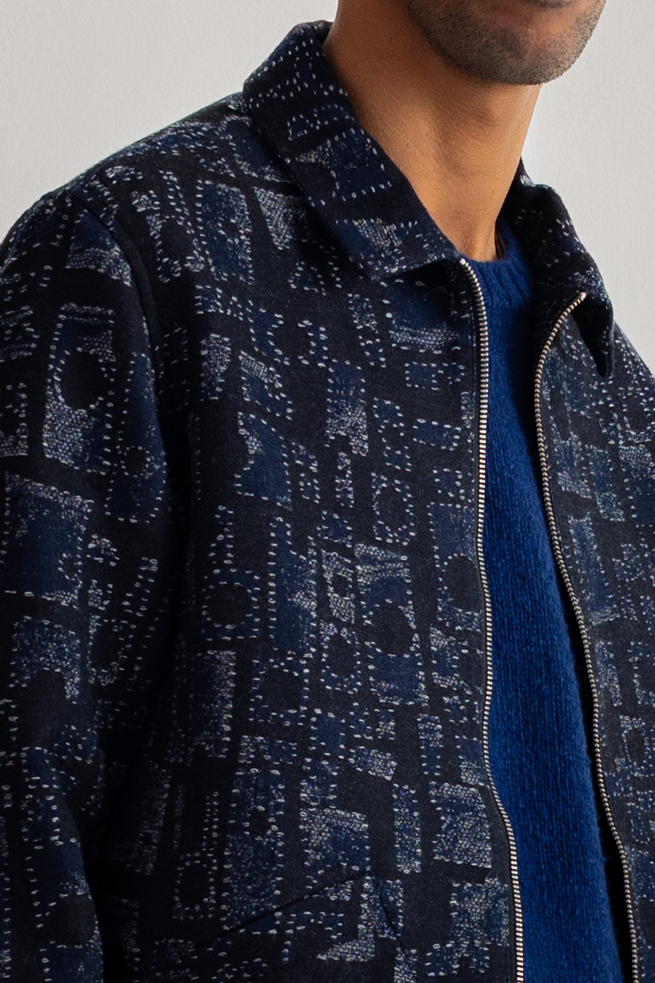 Close-up of a man wearing a dark navy patterned zip-up jacket over a blue knit sweater, against a light gray background.