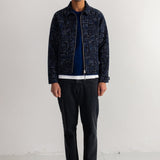 A man stands in a studio wearing a dark patterned zip-up jacket over a navy top, black pants, and black shoes.