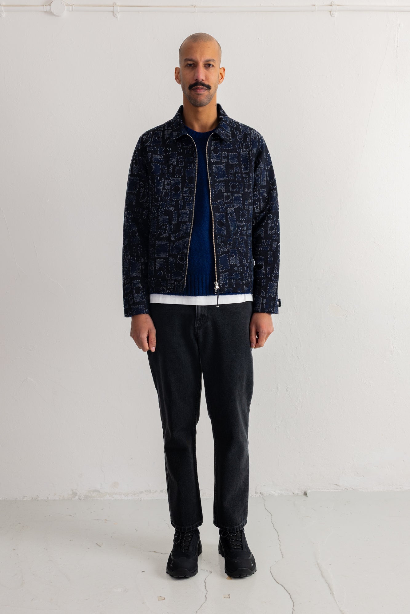 A man stands in a studio wearing a dark patterned zip-up jacket over a navy top, black pants, and black shoes.