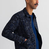 A man wearing a dark navy blue patterned zip-up jacket over a blue sweater and white t-shirt, standing against a white wall.