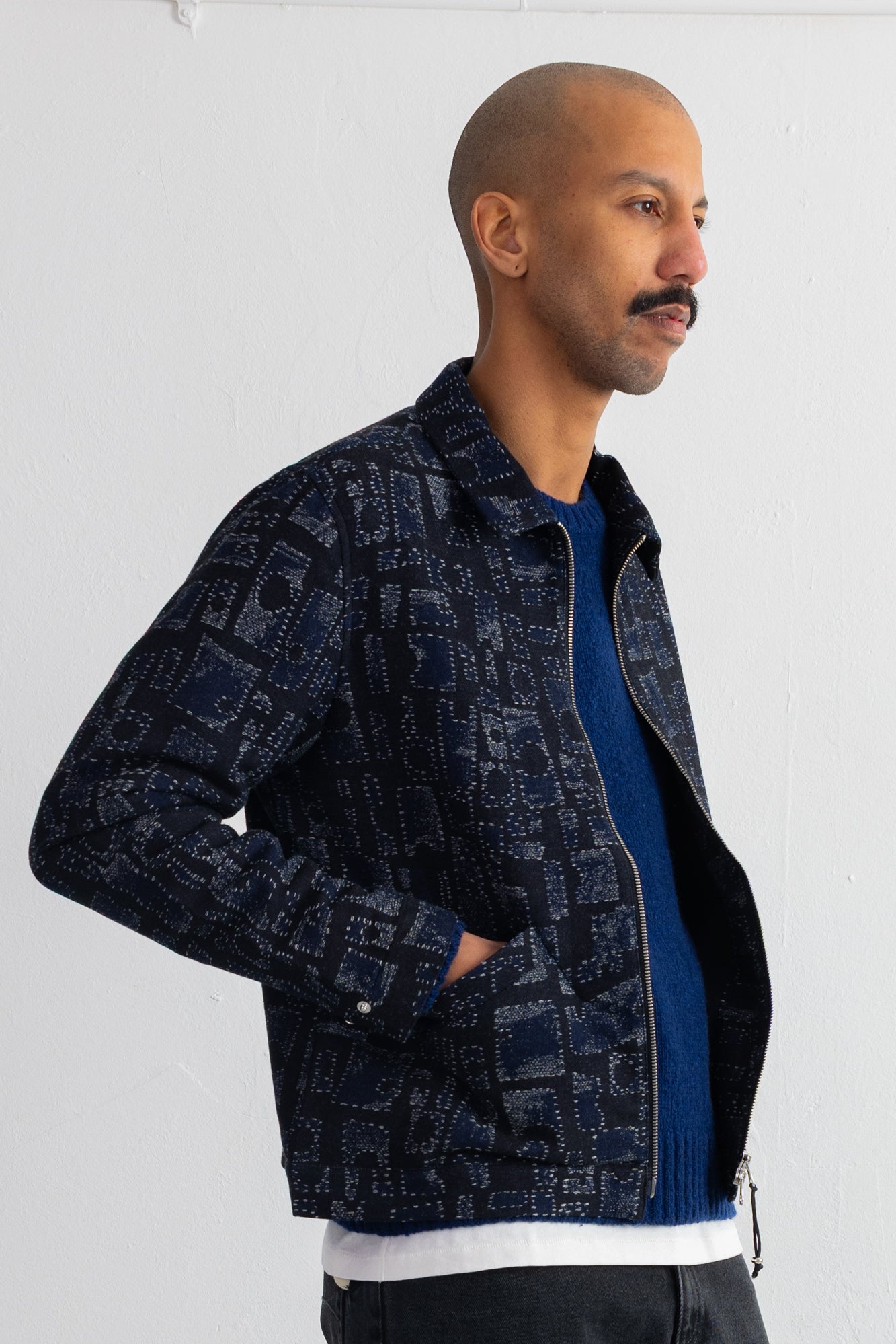 A man wearing a dark navy blue patterned zip-up jacket over a blue sweater and white t-shirt, standing against a white wall.