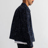 A man shown from the side profile wearing a dark navy blue patterned jacket over a white undershirt and dark jeans, standing against a plain white wall.