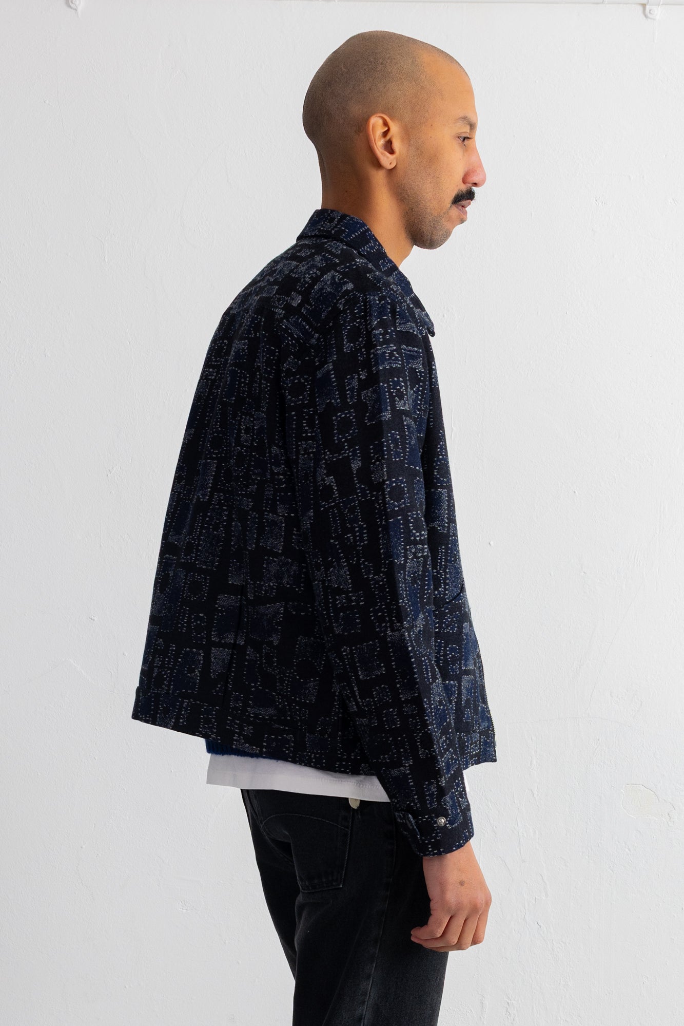A man shown from the side profile wearing a dark navy blue patterned jacket over a white undershirt and dark jeans, standing against a plain white wall.