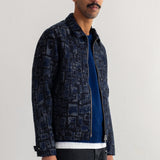 A man wearing a dark navy blue patterned zip-up jacket over a blue sweater and dark jeans, standing against a plain white wall.