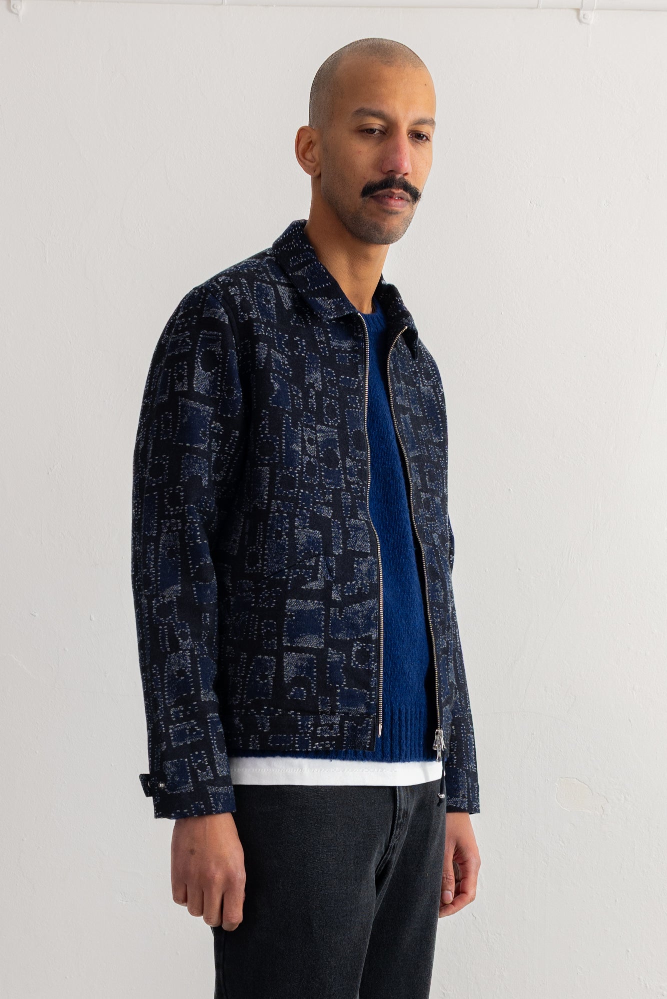 A man wearing a dark navy blue patterned zip-up jacket over a blue sweater and dark jeans, standing against a plain white wall.