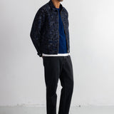 A man wearing a dark patterned bomber jacket over a blue shirt, black pants, and black sneakers, standing against a white studio wall.
